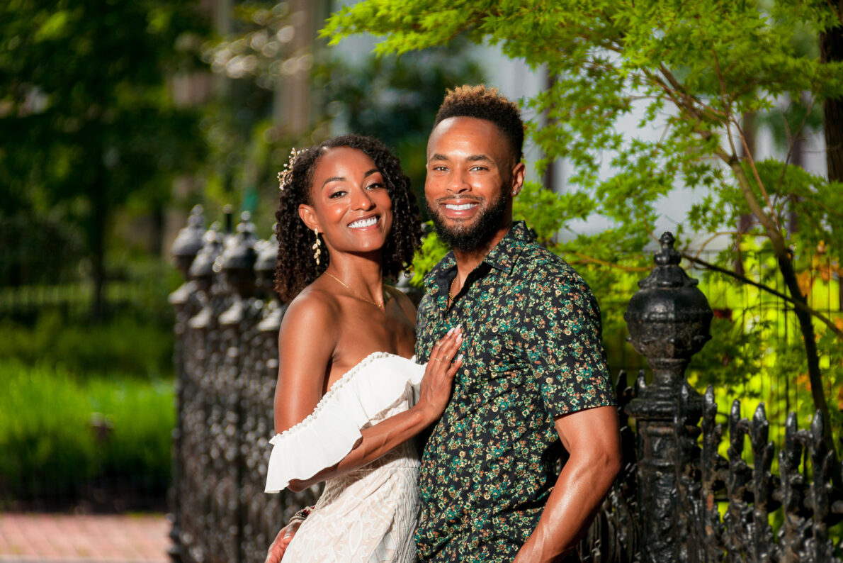 A smiling couple poses in front of a historic wrought iron fence with lush greenery.