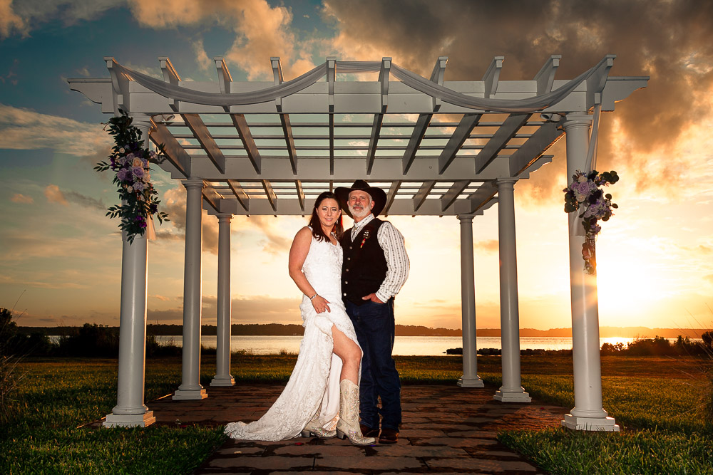 A newlywed couple poses under a floral wedding arch at sunset, with the scenic lChesapeake Bay in the background.